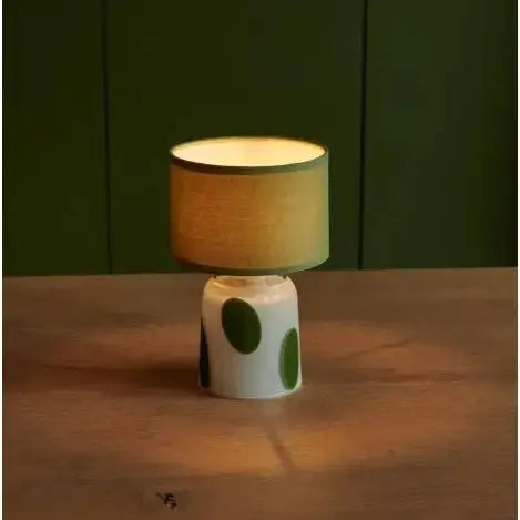 Small lamp with a green and white base on a wooden surface against a dark green wall.