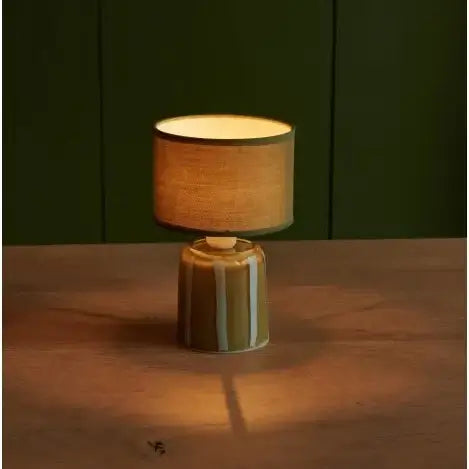 Table lamp with a textured shade on a wooden surface against a dark green wall.
