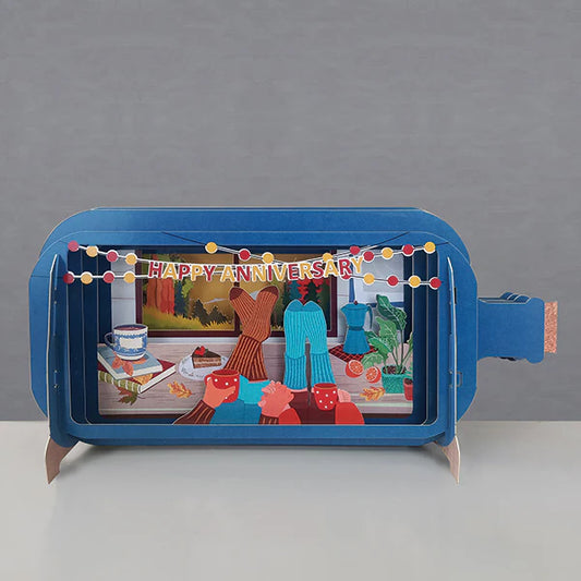 Pop up card in the shape of a bottle with the cosy scene of a couples socked feet resting on a table. Bunting Happy Anniversary across the top