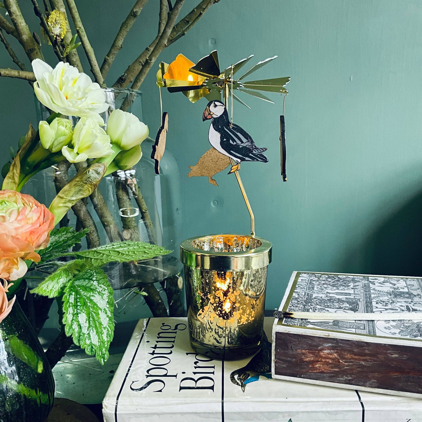 A candle carousel kit with a glass votive and a metal puffin on a table with a floral arrangement and a book in the background.