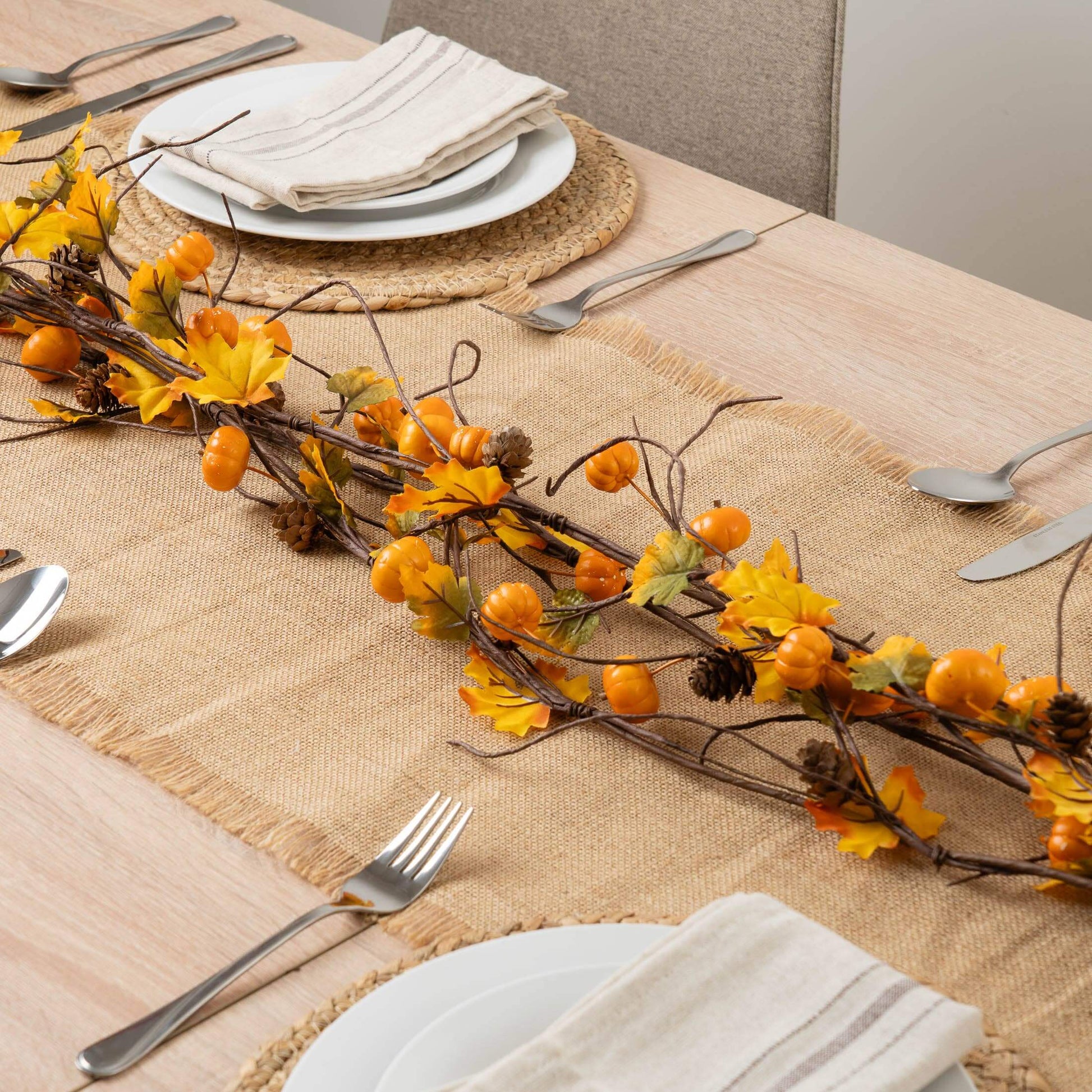 An autumn-themed table runner centerpiece with orange berries, yellow leaves, and pinecones arranged on a woven burlap runner over a light wood dining table set with plates and silverware.