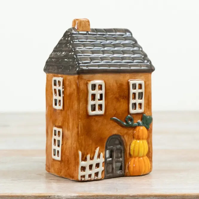 A small decorative ceramic house ornament with a brown body, dark gray roof, white-trimmed windows, a dark door, and orange pumpkins near the front, shown on a light surface against a white background.