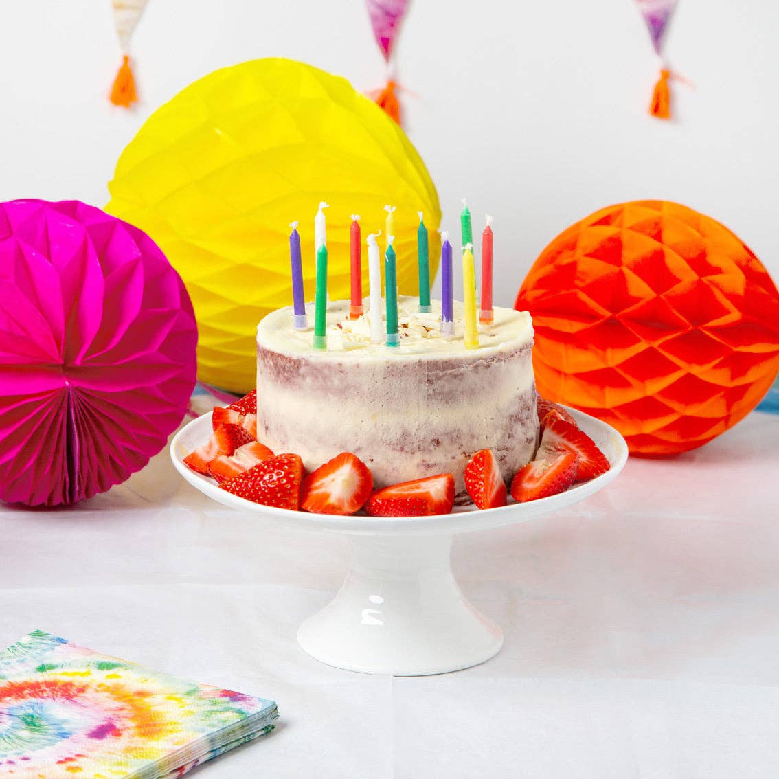 Birthday cake with lit candles on a white plate, surrounded by colorful paper decorations.