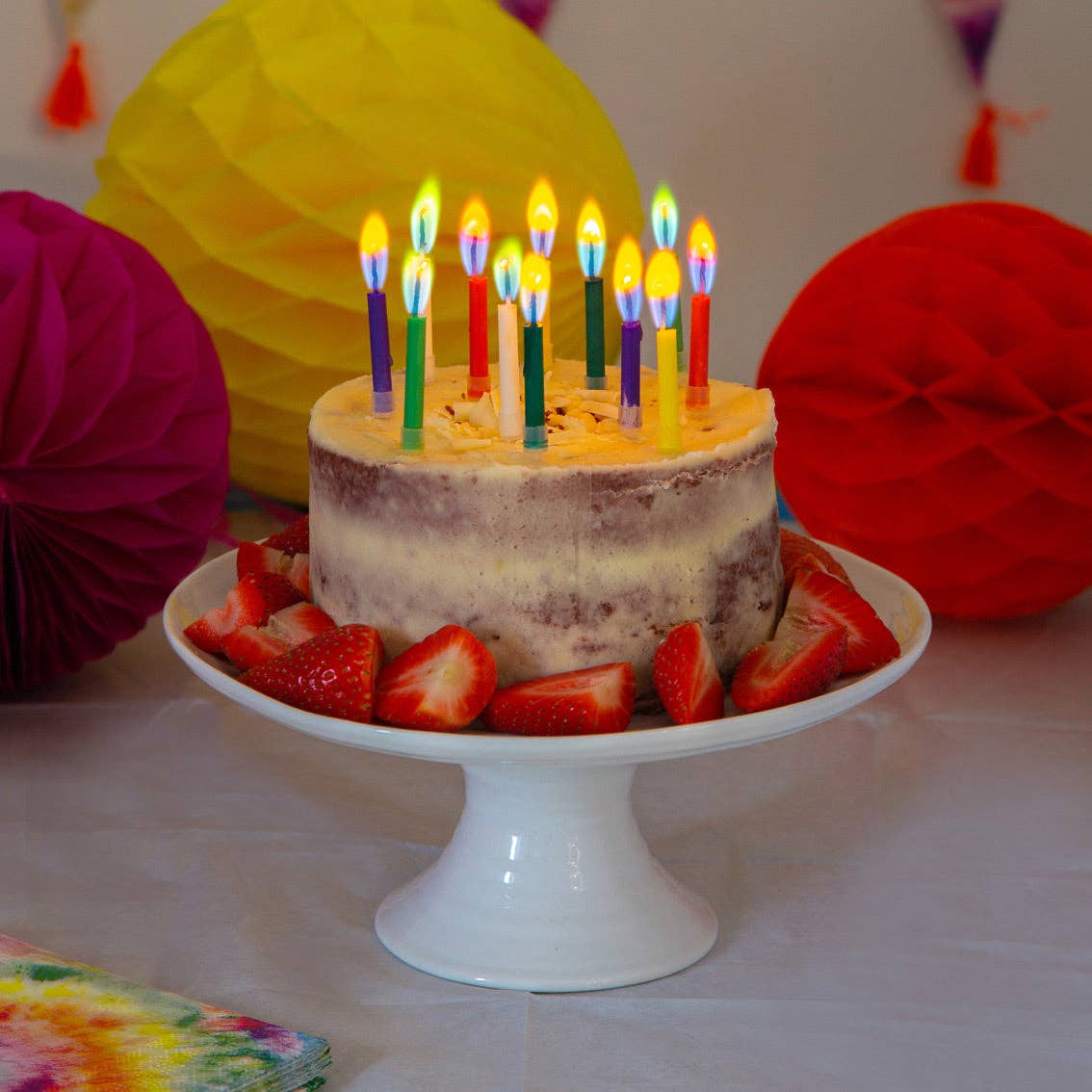 Birthday cake with lit candles on a white plate, surrounded by colorful decorations.
