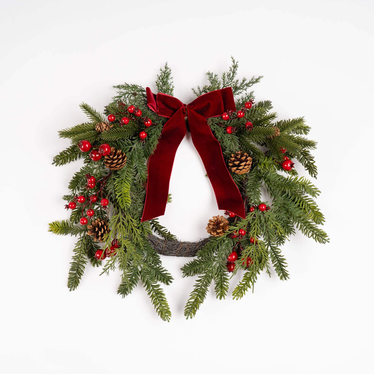 A festive Christmas wreath with green pine branches, red berries, pinecones, and a large red velvet bow, photographed against a plain white background.