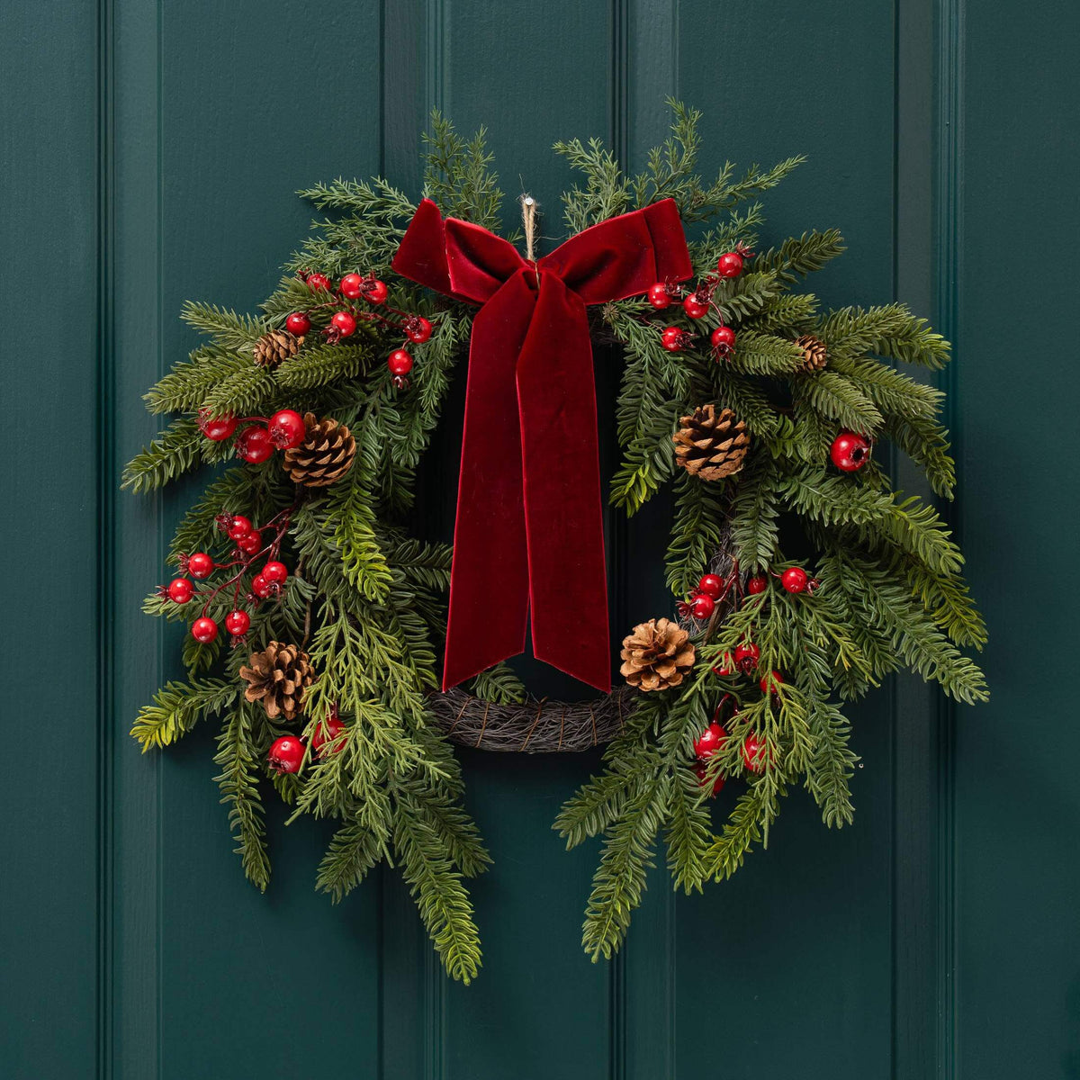 A festive Christmas wreath with green pine branches, red berries, pinecones, and a large red velvet bow, hanging on a dark teal paneled door.