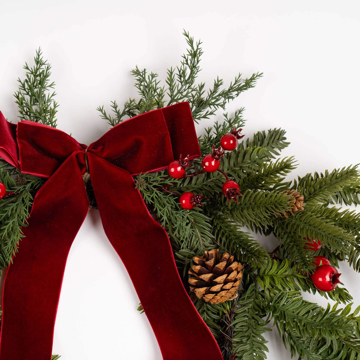 A close-up of a Christmas wreath or garland with green pine branches, a large deep red velvet bow, red berries, and a pinecone on a plain white background.