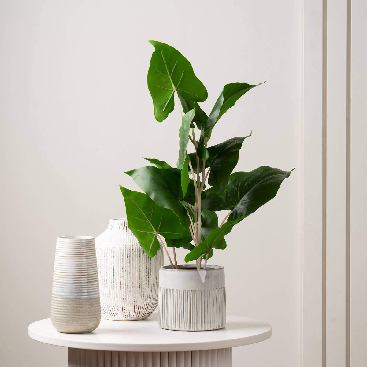 A potted green houseplant with large glossy leaves in a ribbed white planter, styled on a round white side table beside two ceramic vases against a light neutral wall.