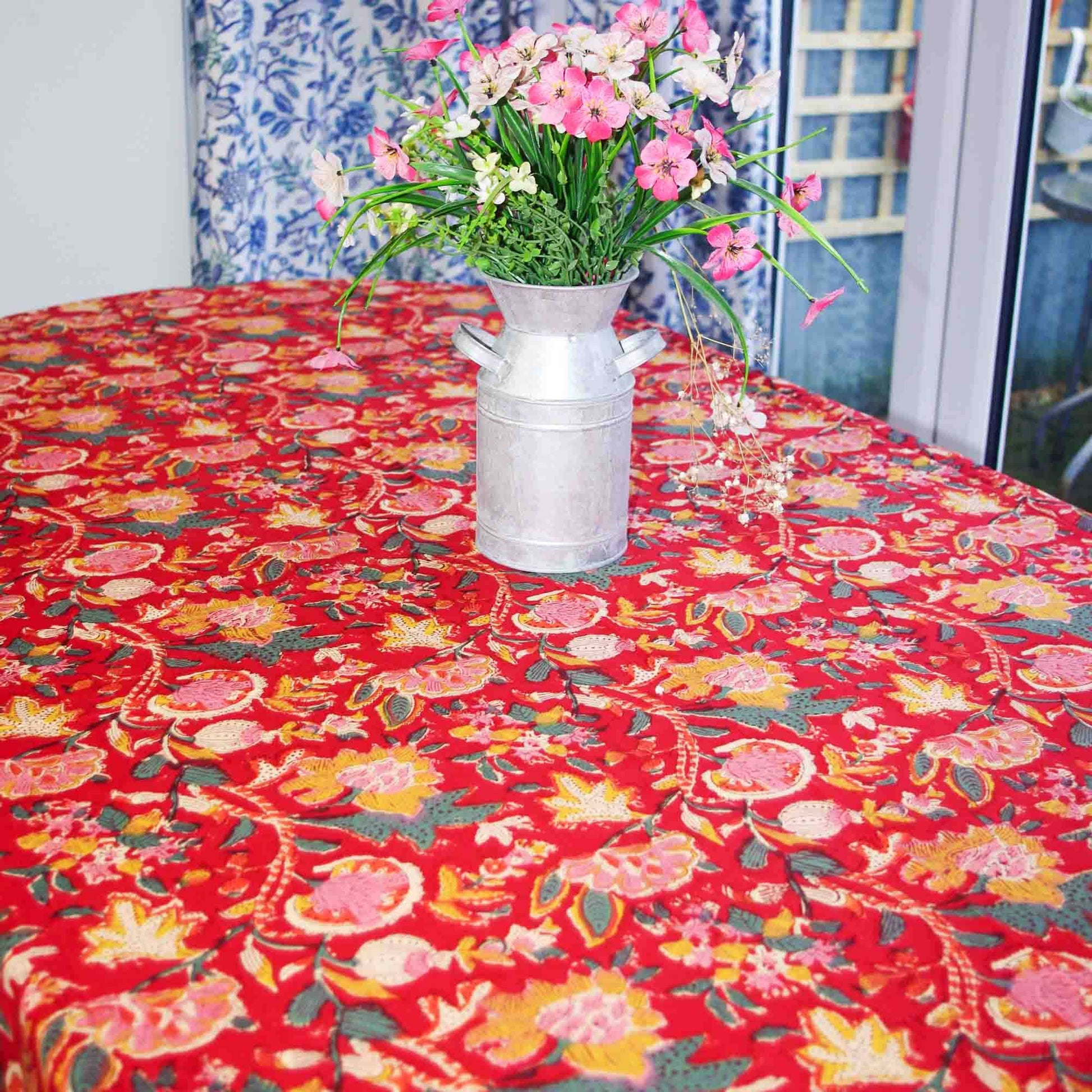 Round table with a red floral tablecloth, silver vase with flowers, and blue patterned curtain in the background.