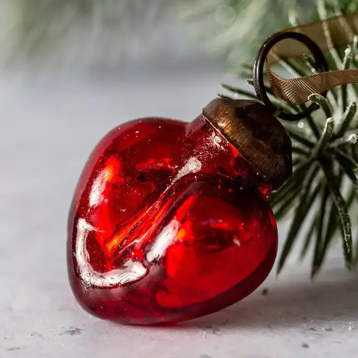 A red heart-shaped glass Christmas ornament with a metallic bronze cap and a brown ribbon hanger, resting on a light gray surface with blurred green pine needles in the background.