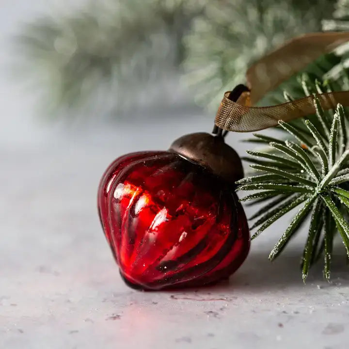 A red ribbed glass Christmas ornament with a brown cap and a brown ribbon loop, resting on a light gray surface next to green frosted pine branches.