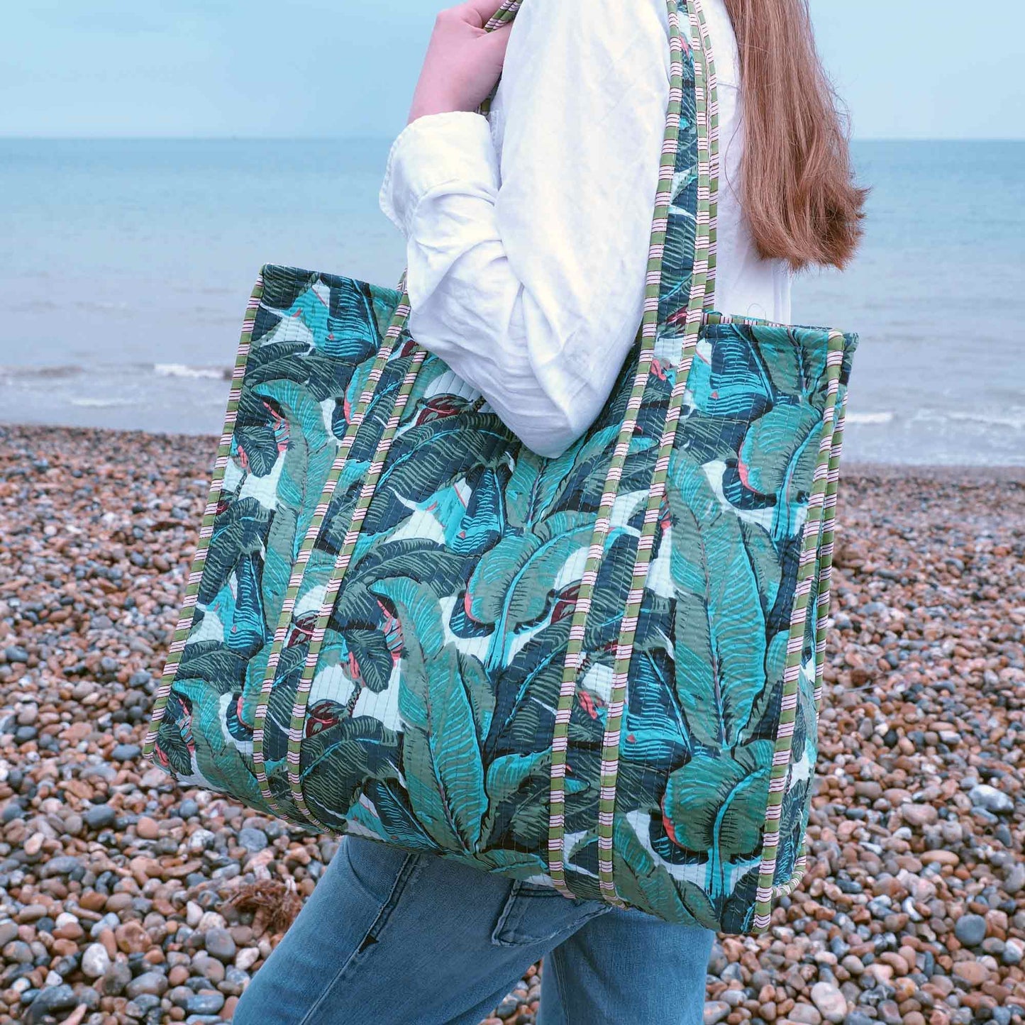 Person holding a green leaf-patterned tote bag on a pebbly beach.