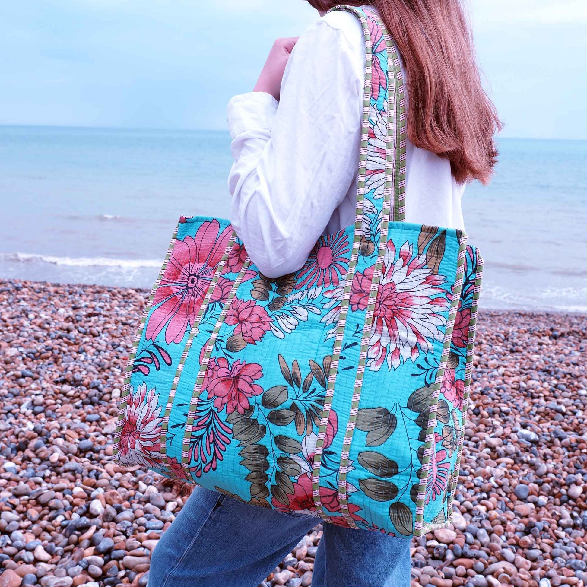 Person holding a floral tote bag on a pebbly beach with ocean in the background