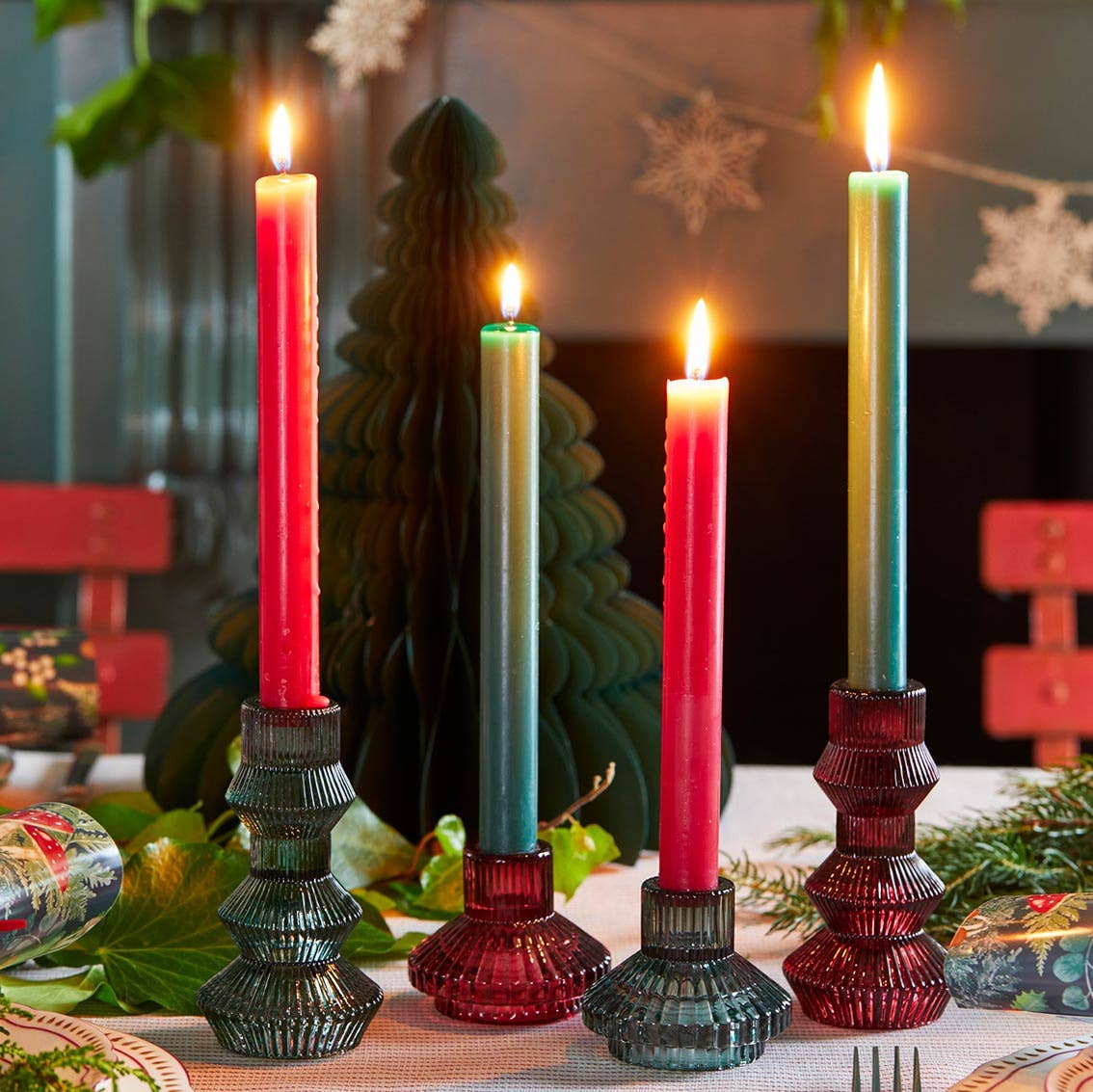 Set of lit candles in decorative holders on a table with a Christmas tree and decorations in the background.
