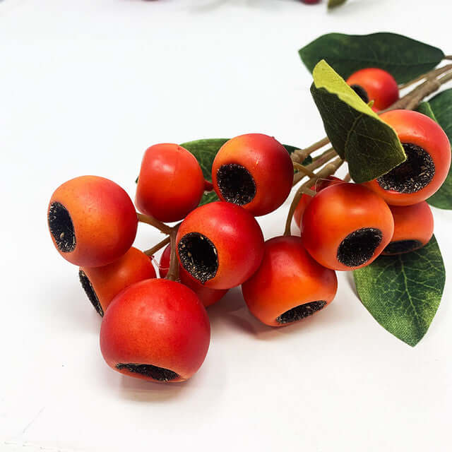 Artificial red rose hips on a white background, featuring realistic red berries with hollow black centers and green leaves.