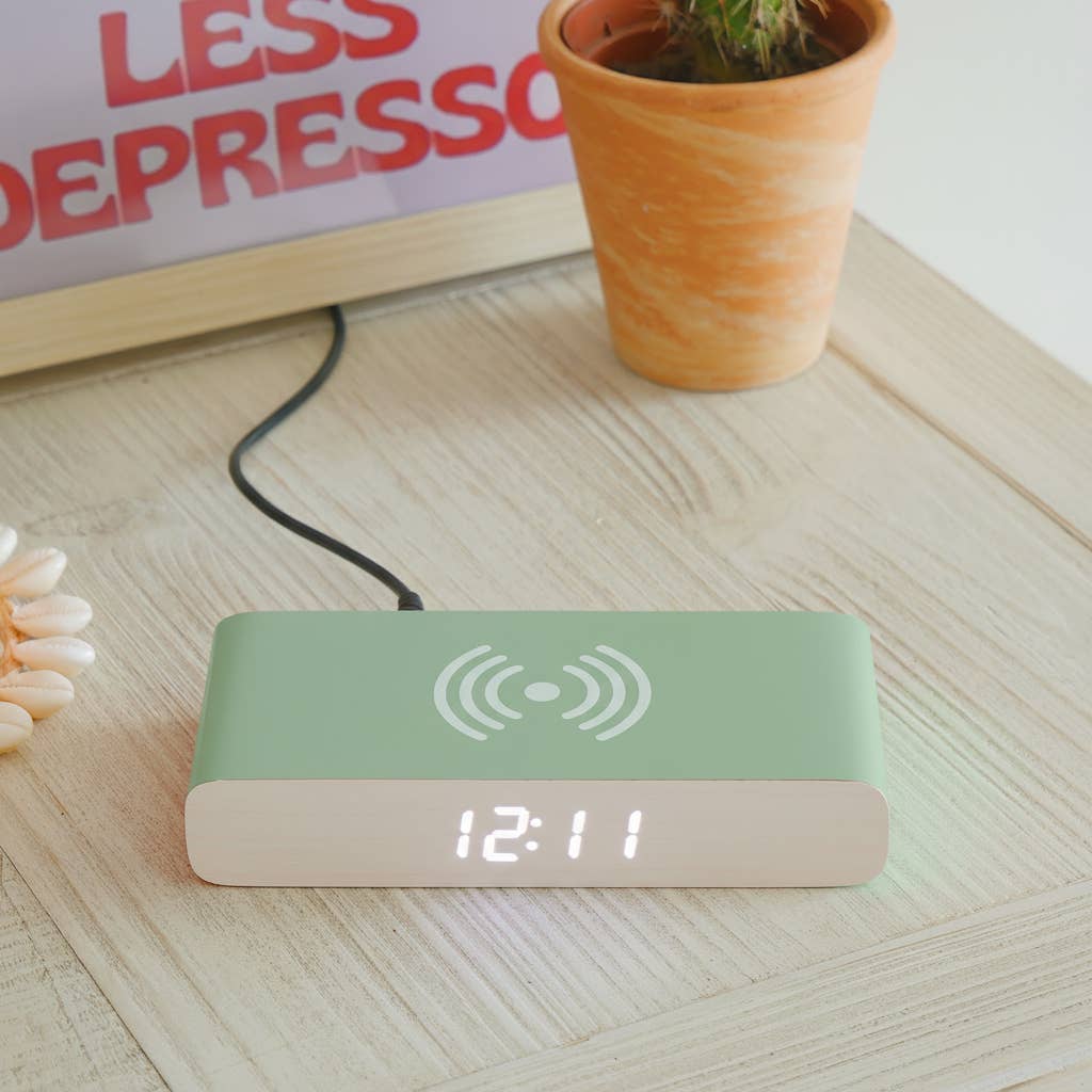 Green digital clock with wooden base on a light surface, next to a potted plant and motivational poster.