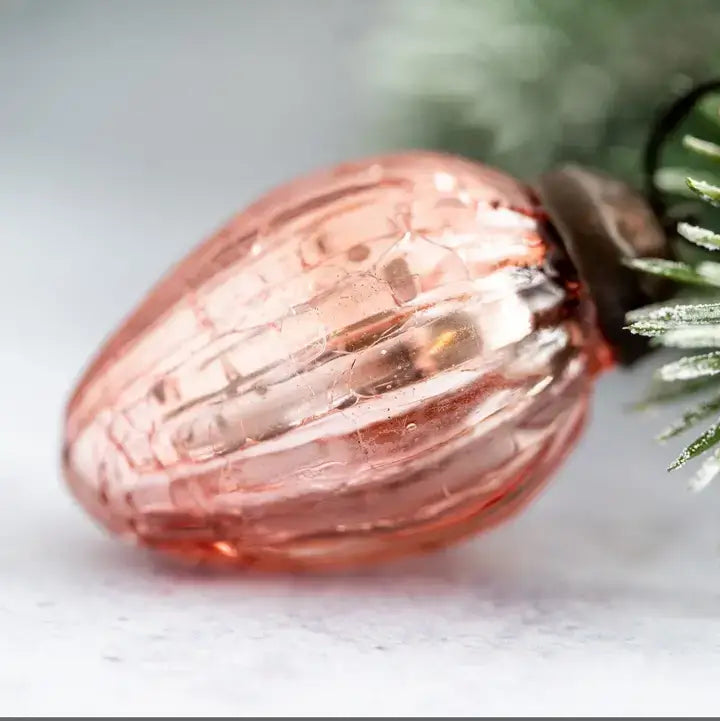 A close-up of a ribbed, translucent pink glass ornament shaped like a pinecone, resting on a light gray surface with blurred green pine needles in the background.