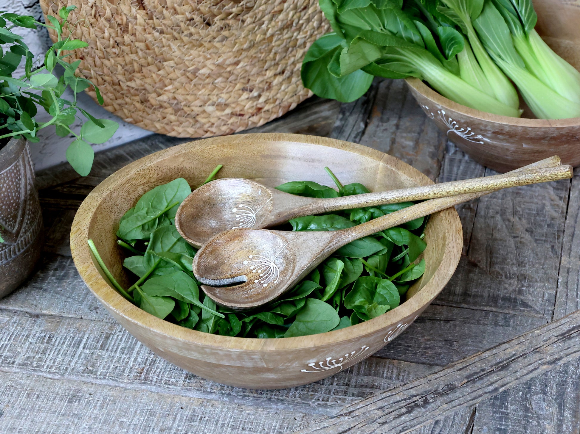 Wooden bowl with green leafy vegetables and wooden spoons on a rustic wooden surface.