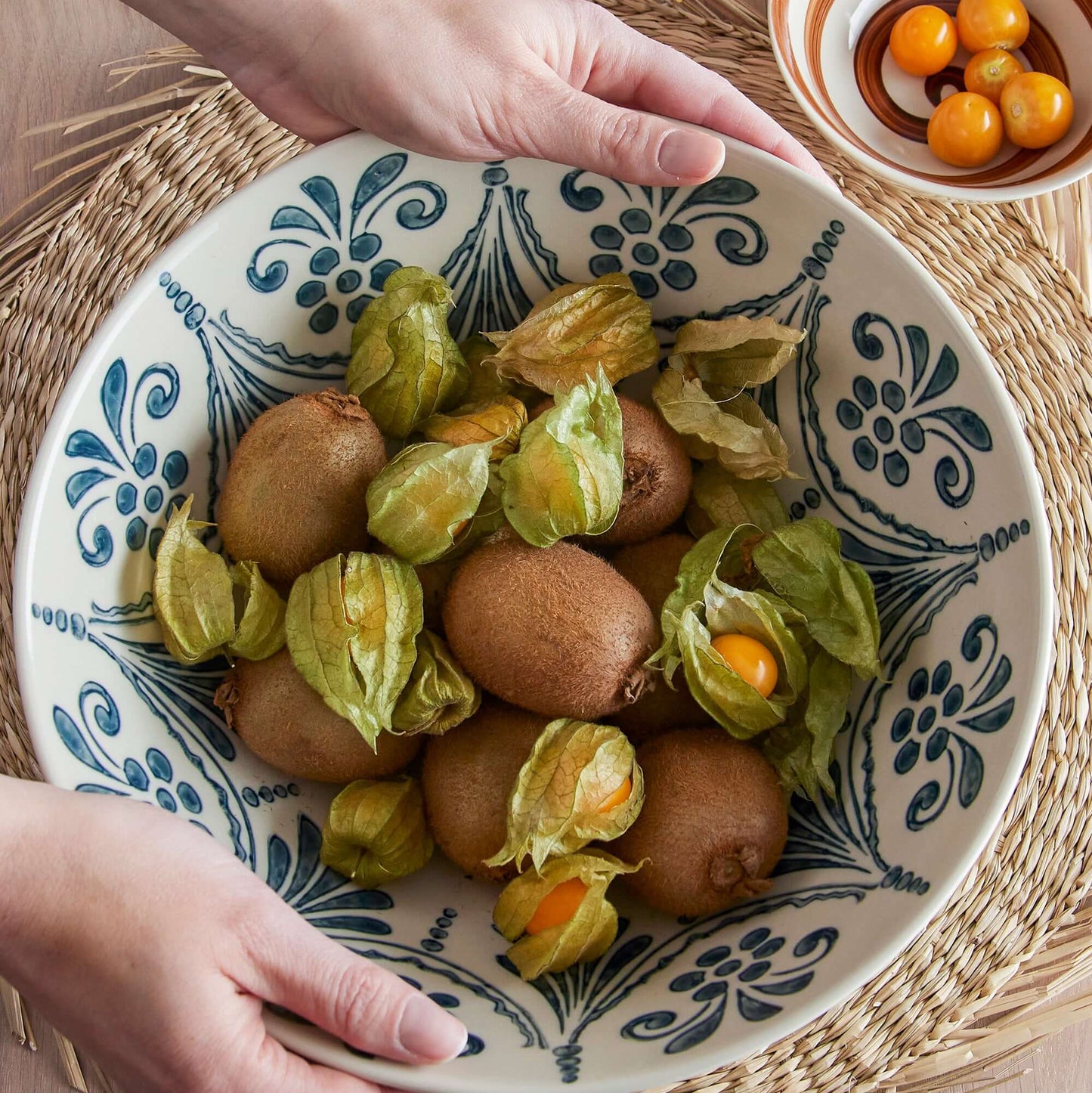Person holding a decorative bowl with physalis and eggs on a woven mat.