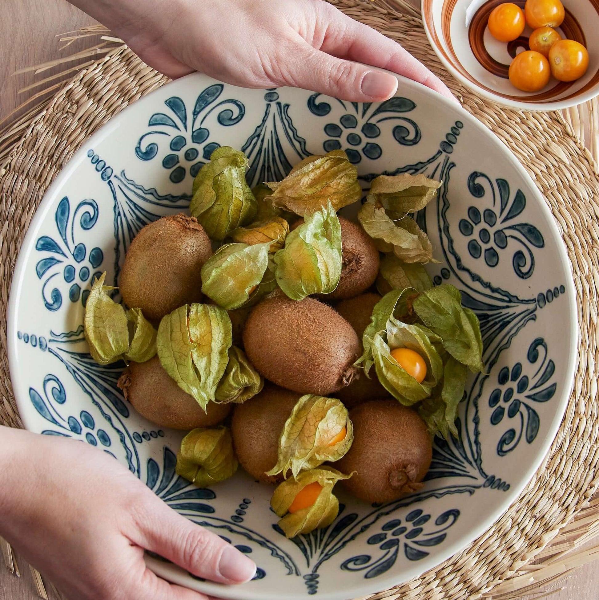 Person holding a decorative bowl with physalis and eggs on a woven mat.
