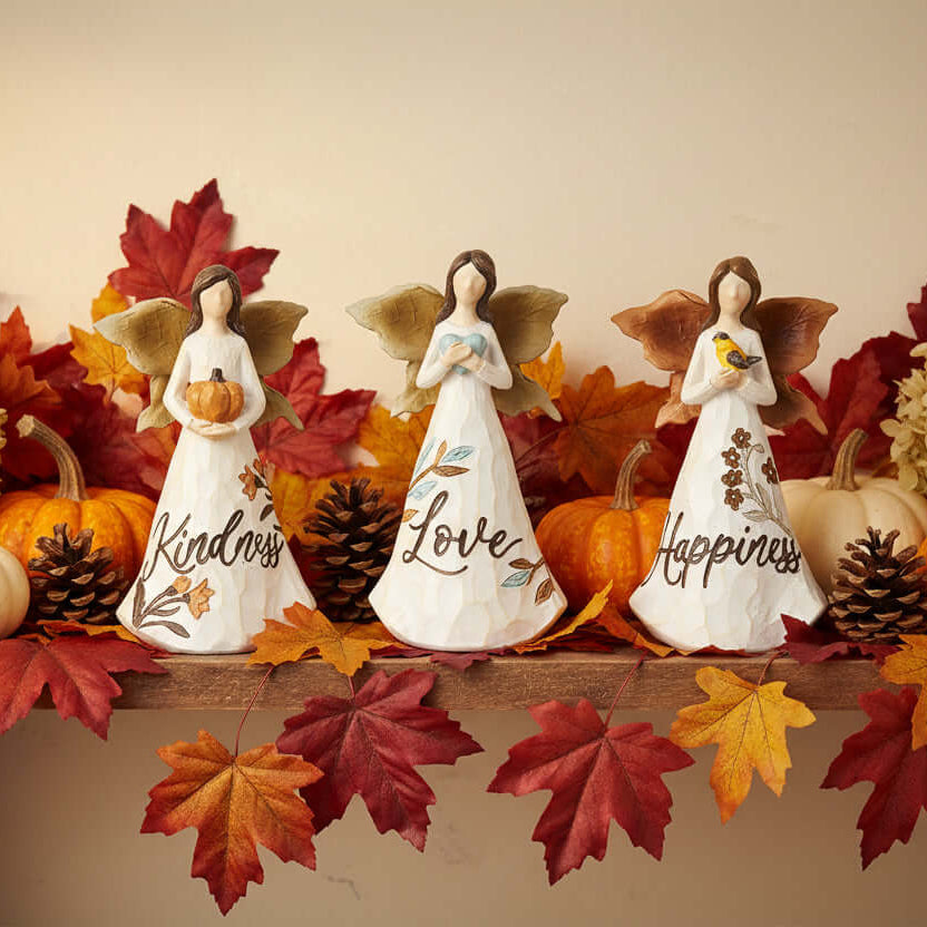Three angel figurines with wings, each holding a symbol and word on a shelf with autumn leaves and pumpkins