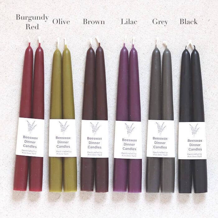 Set of six different colored candles labeled Burgundy, Olive, Brown, Lilac, Grey, and Black on a textured white background.