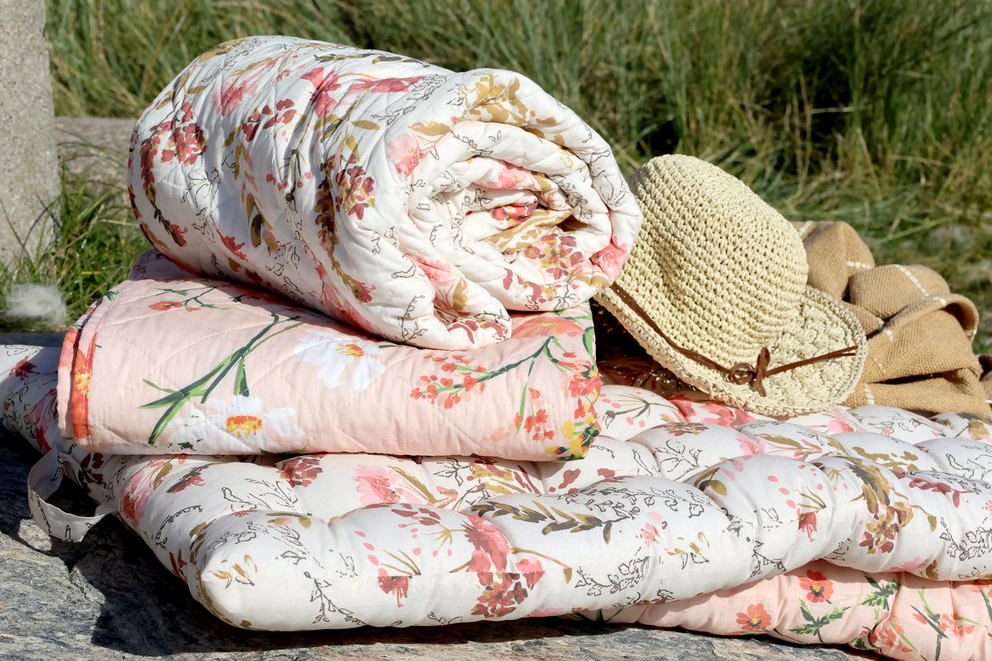 Stack of floral-patterned quilts with a straw hat and coat on a grassy background