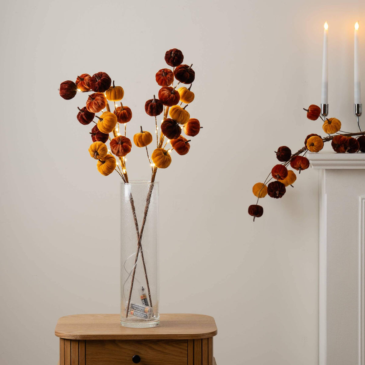 Autumn pumpkin stem arrangement with small orange, yellow, and dark red pumpkins in a clear glass vase on a wooden side table, with a matching branch draped over a white mantel with lit white taper candles.