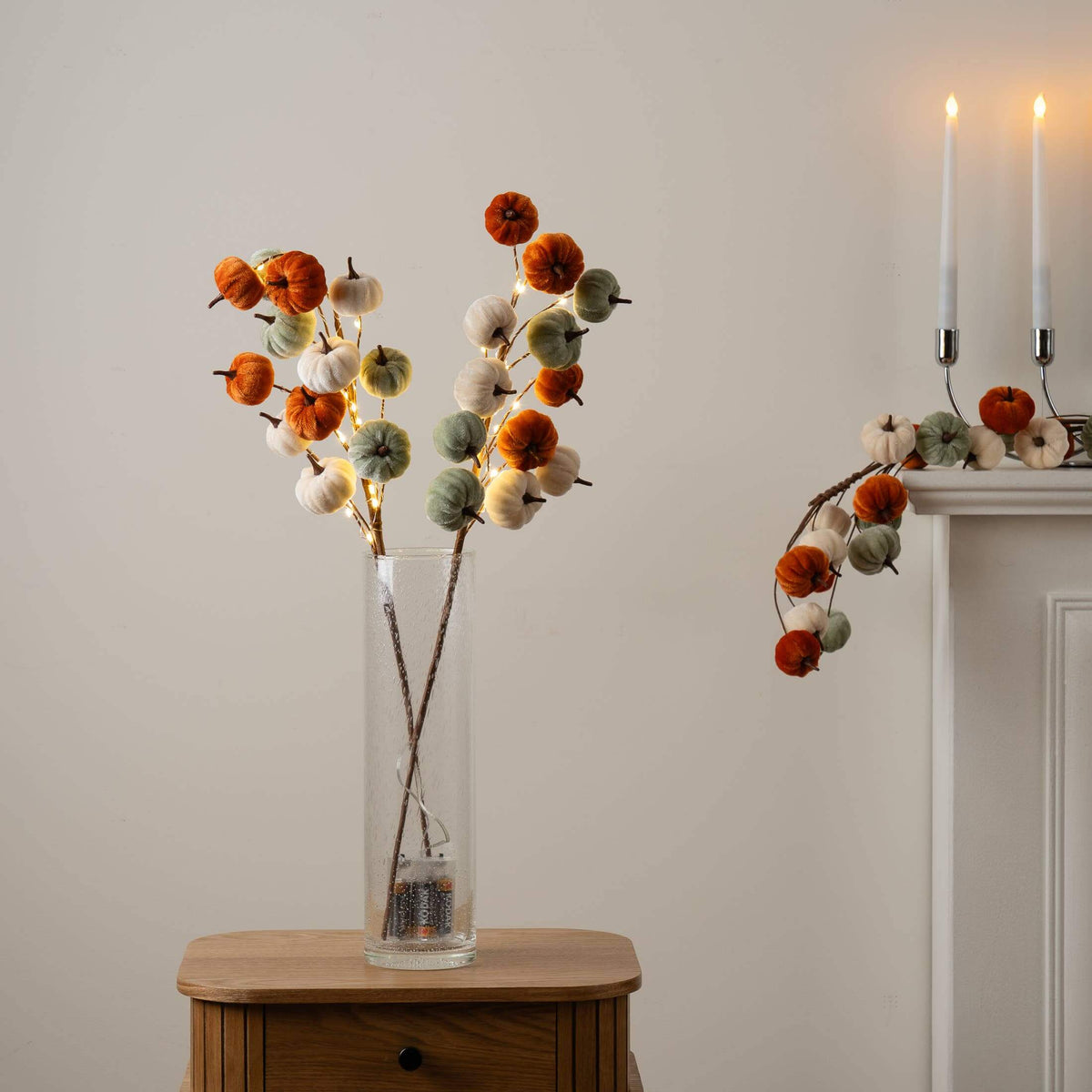 Autumn-themed artificial pumpkin garland with warm white fairy lights, featuring orange, sage green, and cream cotton bolls on brown stems, displayed in a tall clear glass vase on a wooden side table and draped over a white mantel with lit taper candles.
