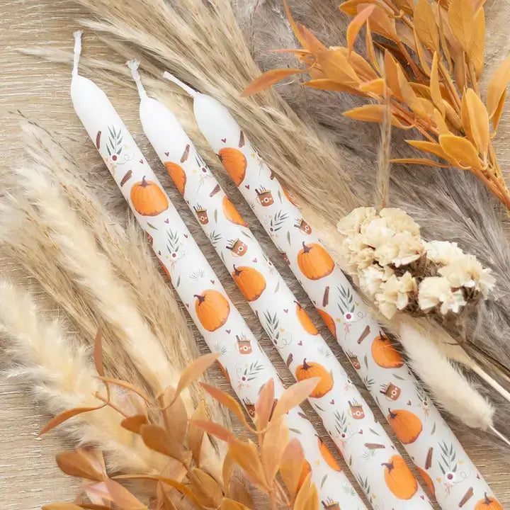 Three white candles with autumn-themed designs on a wooden surface with decorative elements.