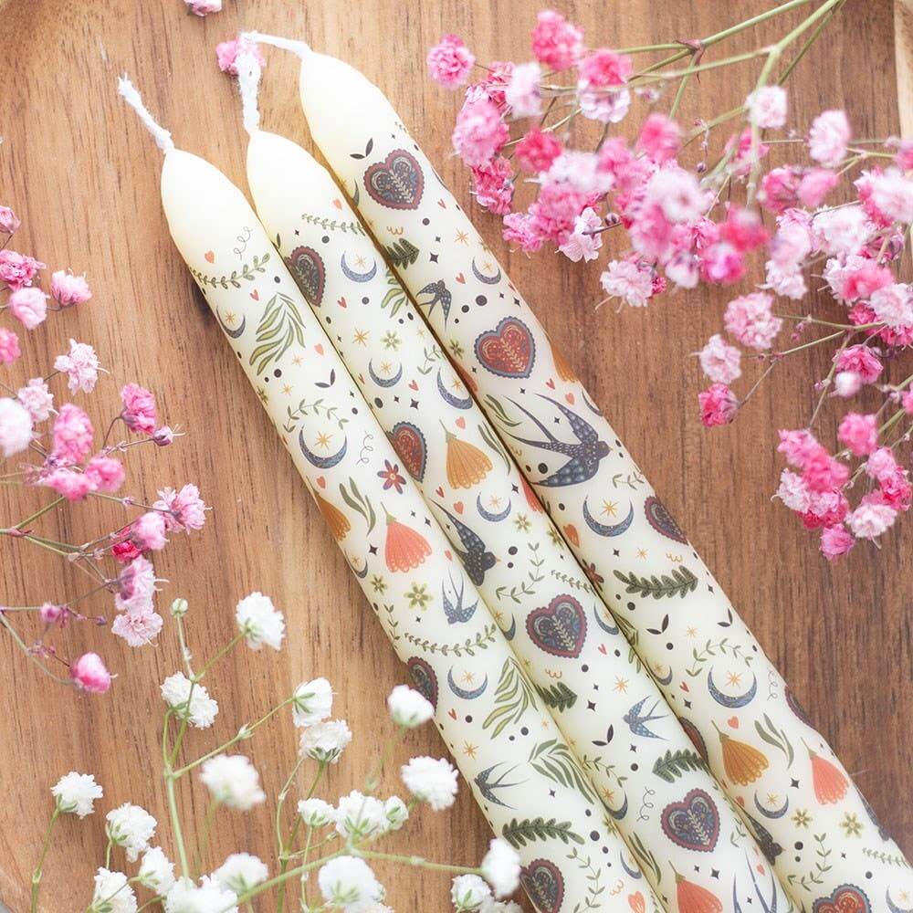 Decorative candles with heart and moon patterns on a wooden surface with pink and white flowers.