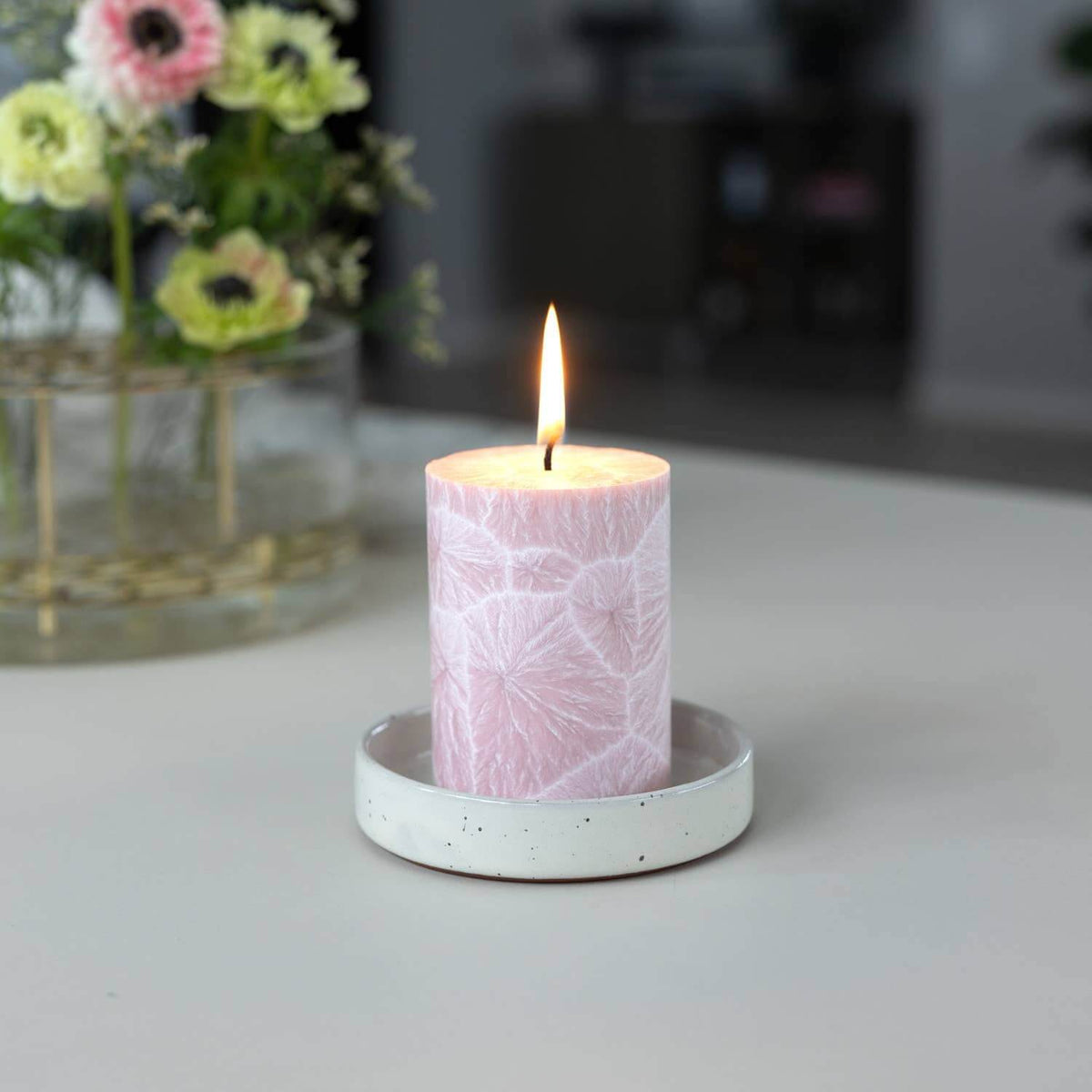 A lit pink candle with a white floral pattern sits in a small speckled white ceramic holder on a tabletop, with a blurred vase of flowers in the background.