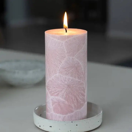 A lit pink pillar candle with a textured leaf pattern sits in a speckled white ceramic holder on a light gray tabletop.