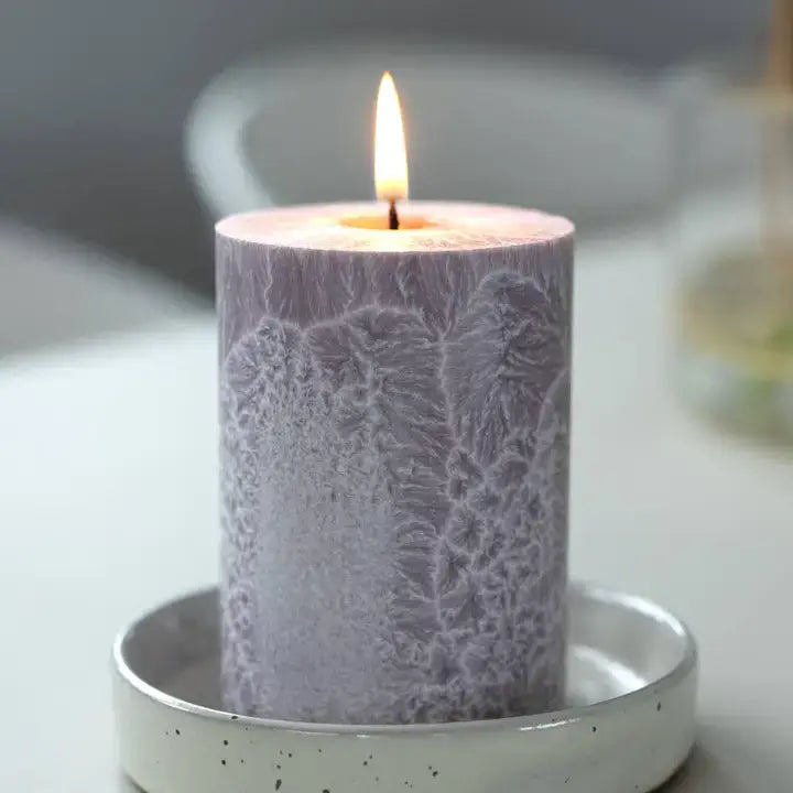 A lit lavender pillar candle with a textured, frosty pattern sits in a speckled white ceramic holder on a light tabletop.