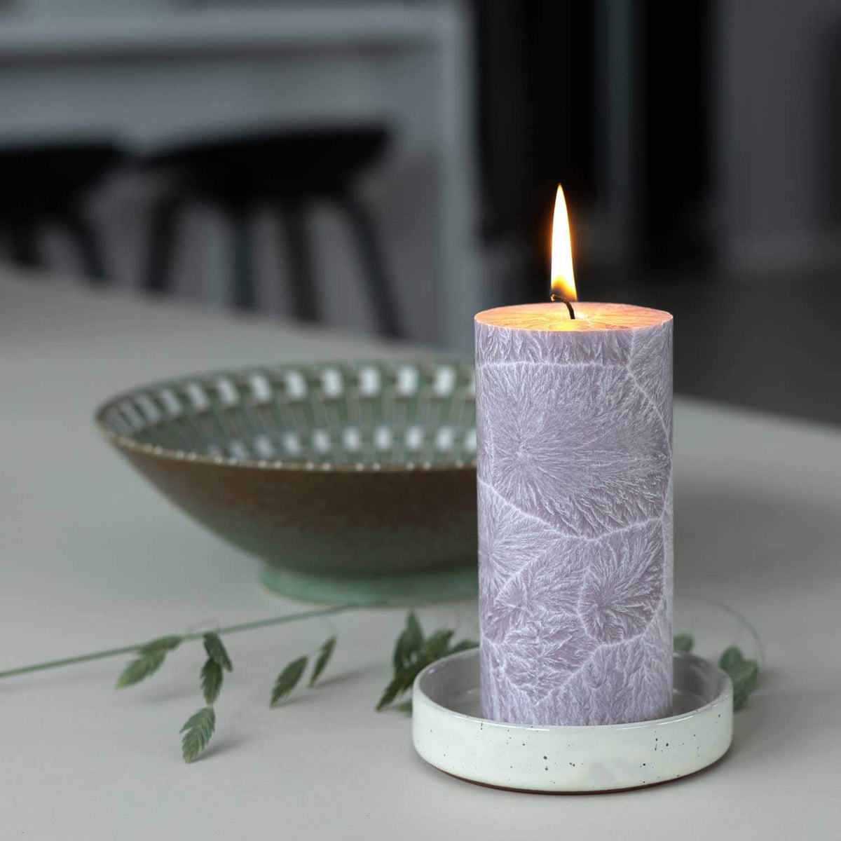 A lit cylindrical candle with a pale lavender floral pattern sits on a white ceramic holder on a light tabletop. A blurred bowl and greenery are in the background.