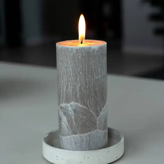 A lit gray pillar candle with a textured, leaf-like pattern sits in a white speckled ceramic holder on a neutral gray surface.