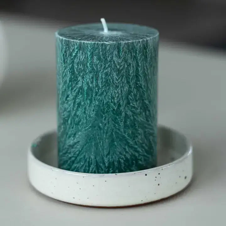 A textured teal green pillar candle with a white wick, sitting in a white speckled ceramic candle holder on a neutral gray surface.