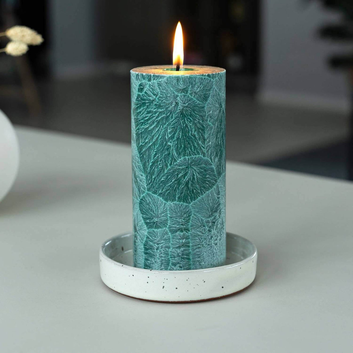 Teal candle with leaf pattern on a white base against a blurred background