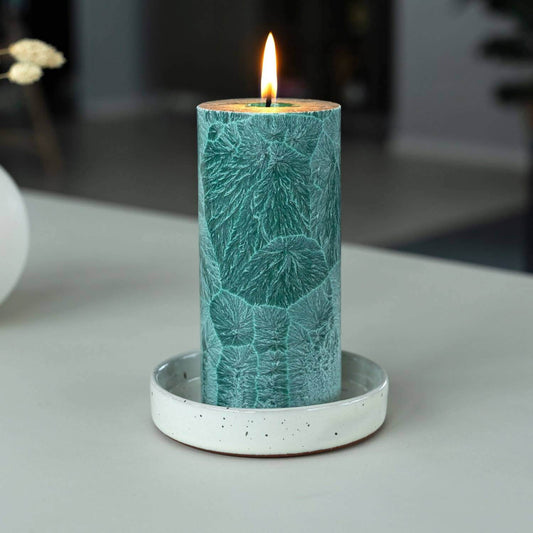 Teal candle with leaf pattern on a white base against a blurred background