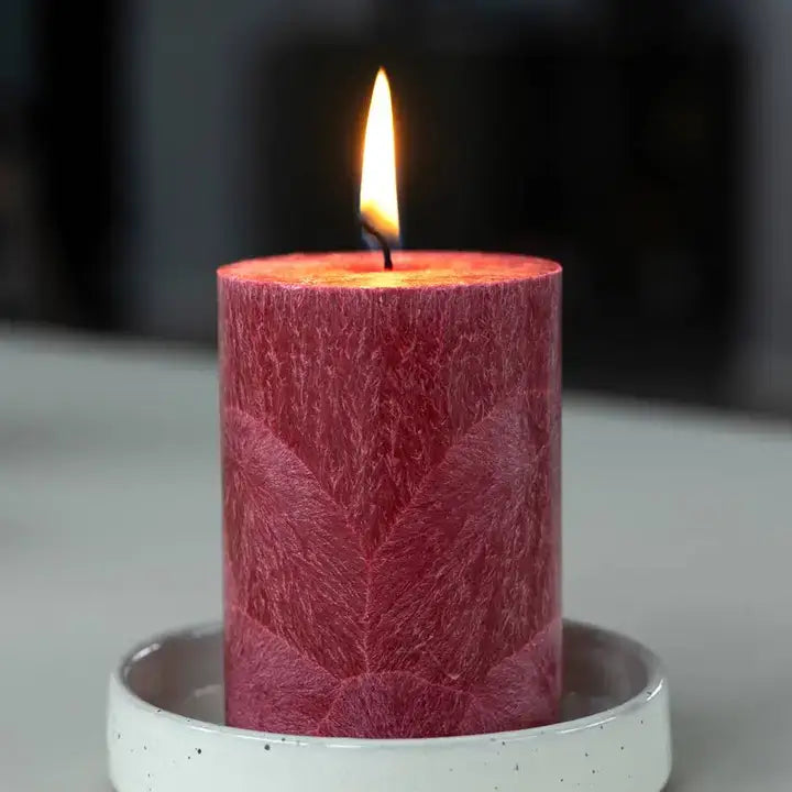 A lit red pillar candle with a textured, leaf-like embossed pattern, sitting in a white speckled ceramic holder against a dark, blurred background.