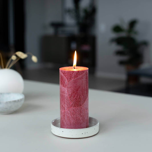 A lit pink pillar candle with a textured, leaf-like pattern sits in a white speckled ceramic holder on a light gray tabletop, with a softly blurred modern interior background.