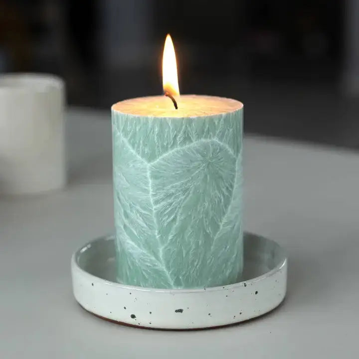 A lit teal-green leaf-patterned candle in a white speckled ceramic holder, set on a gray surface with a blurred background.