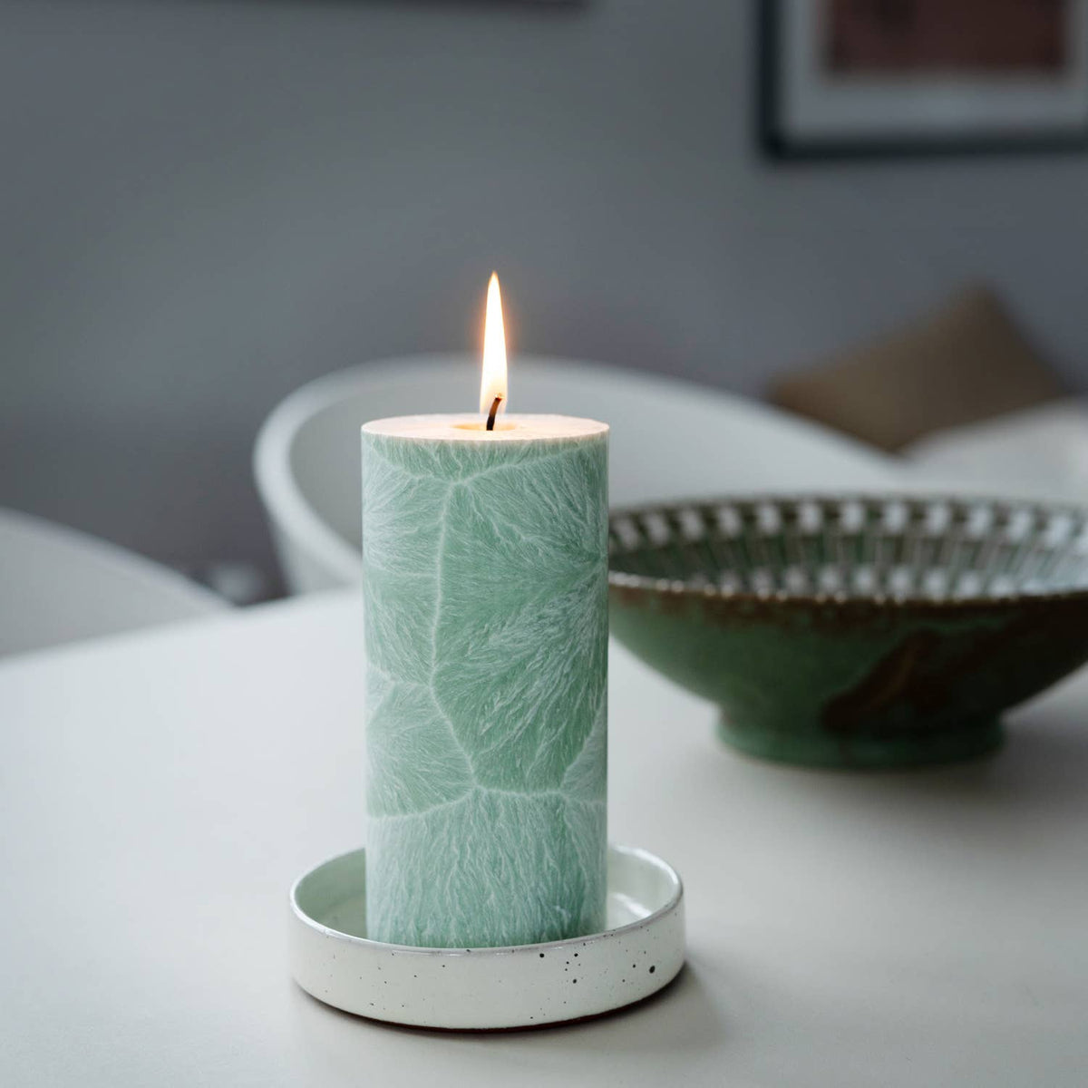 A lit cylindrical candle in a white ceramic holder, featuring a pale mint green leaf pattern, set on a white table with a blurred green bowl in the background.