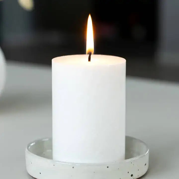 A lit white pillar candle standing in a small speckled white ceramic holder, photographed close up against a dark blurred background.