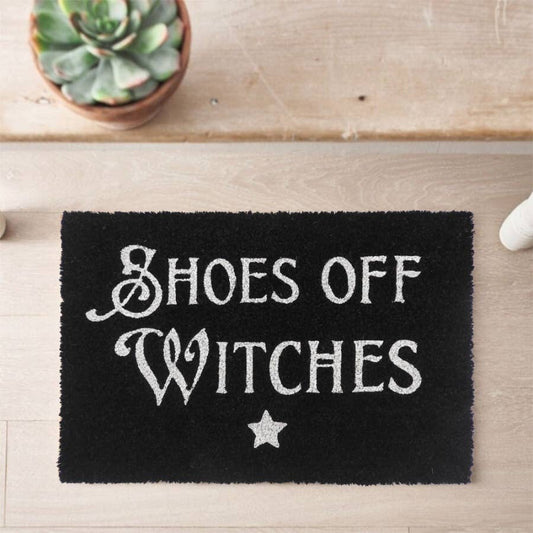 Black doormat with 'Shoes Off Witches' text on a wooden floor.
