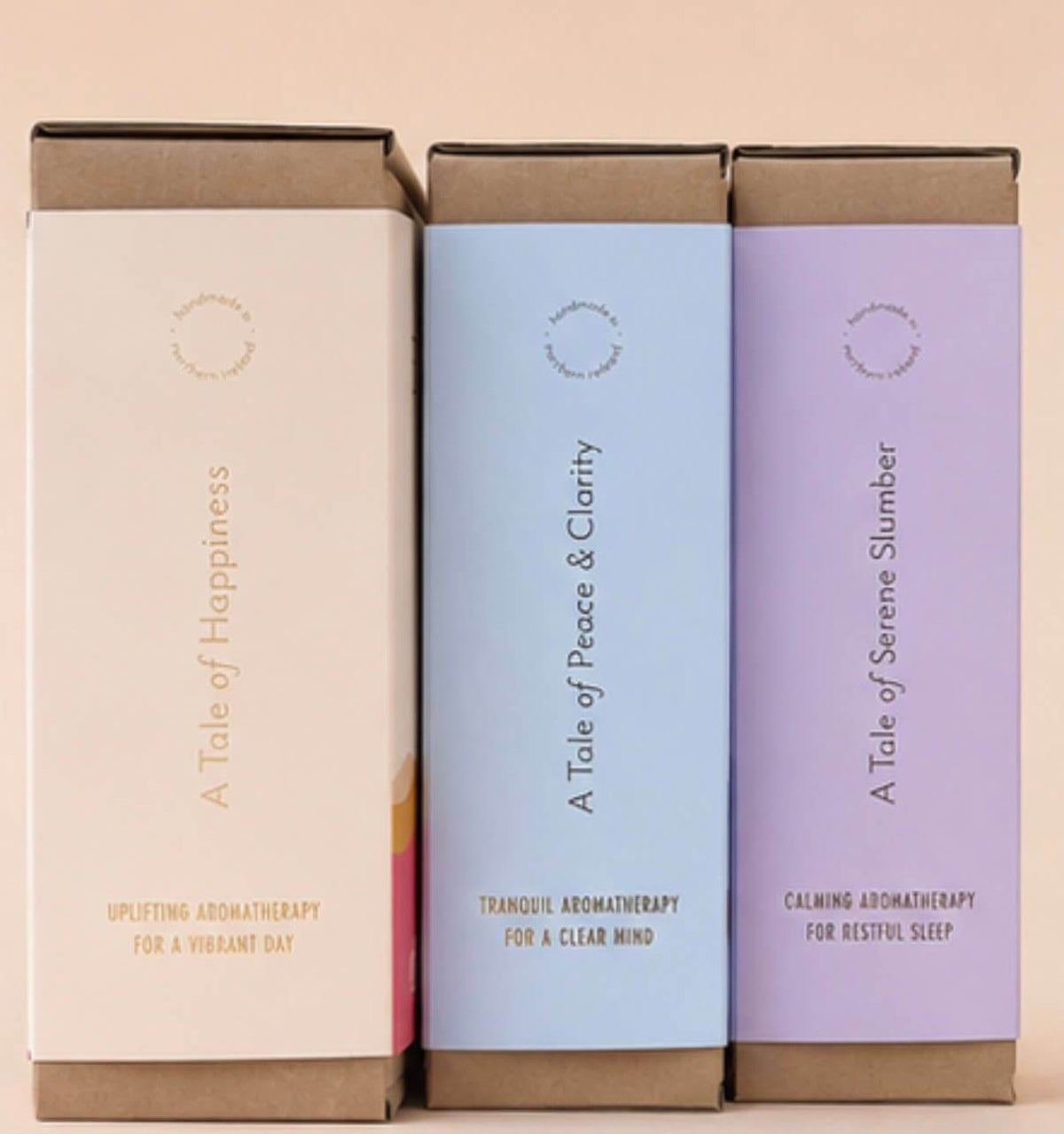 Three rectangular boxed products standing upright side by side on a plain beige background. The boxes have kraft brown top and bottom sections with pastel front panels in cream, light blue, and light purple, each with vertical text and small circular logo details.