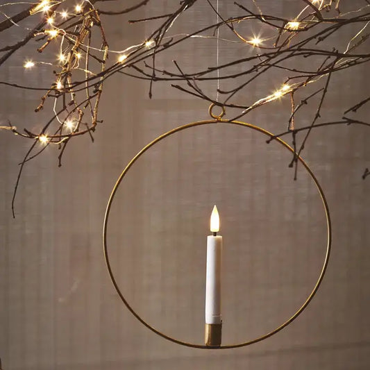 An LED candle ring with a rustic gold finish, hanging from a branch, with a lit candle inside it, surrounded by warm white LED lights.