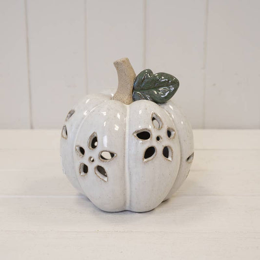 A white ceramic decorative pumpkin with cutout details, a beige stem, and a glossy dark green leaf on a white wooden surface.