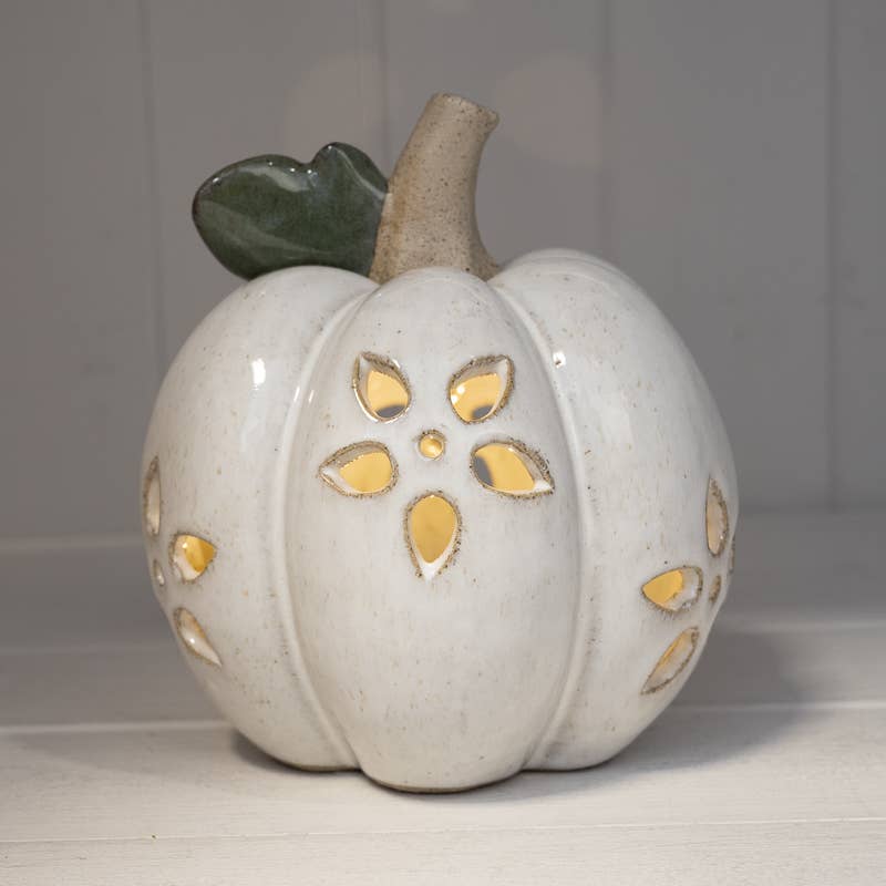 A ceramic decorative pumpkin with a speckled off-white glaze, featuring cutout floral patterns that reveal a warm yellow interior, a textured tan stem, and a dark green leaf on top.