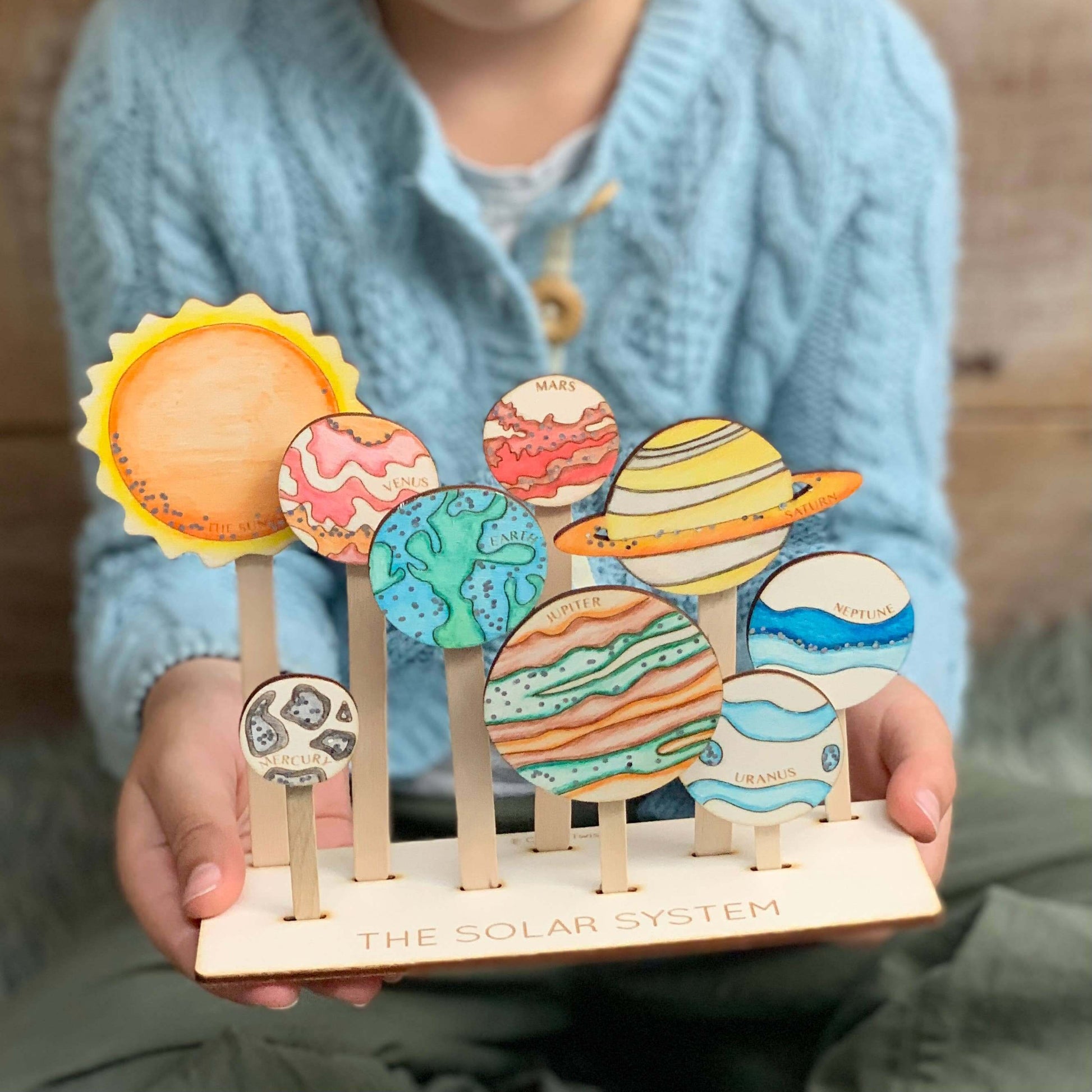 A child holding a craft kit solar system display stand with colorful wooden planets and a sun, completed and presented on a wooden surface.
